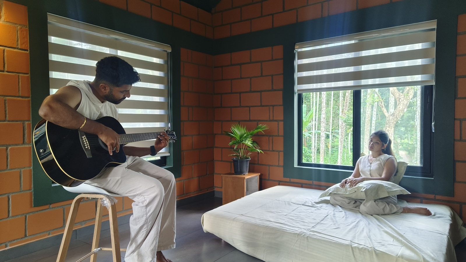 A man playing guitar while a woman relaxes on the bed in a brick-walled room