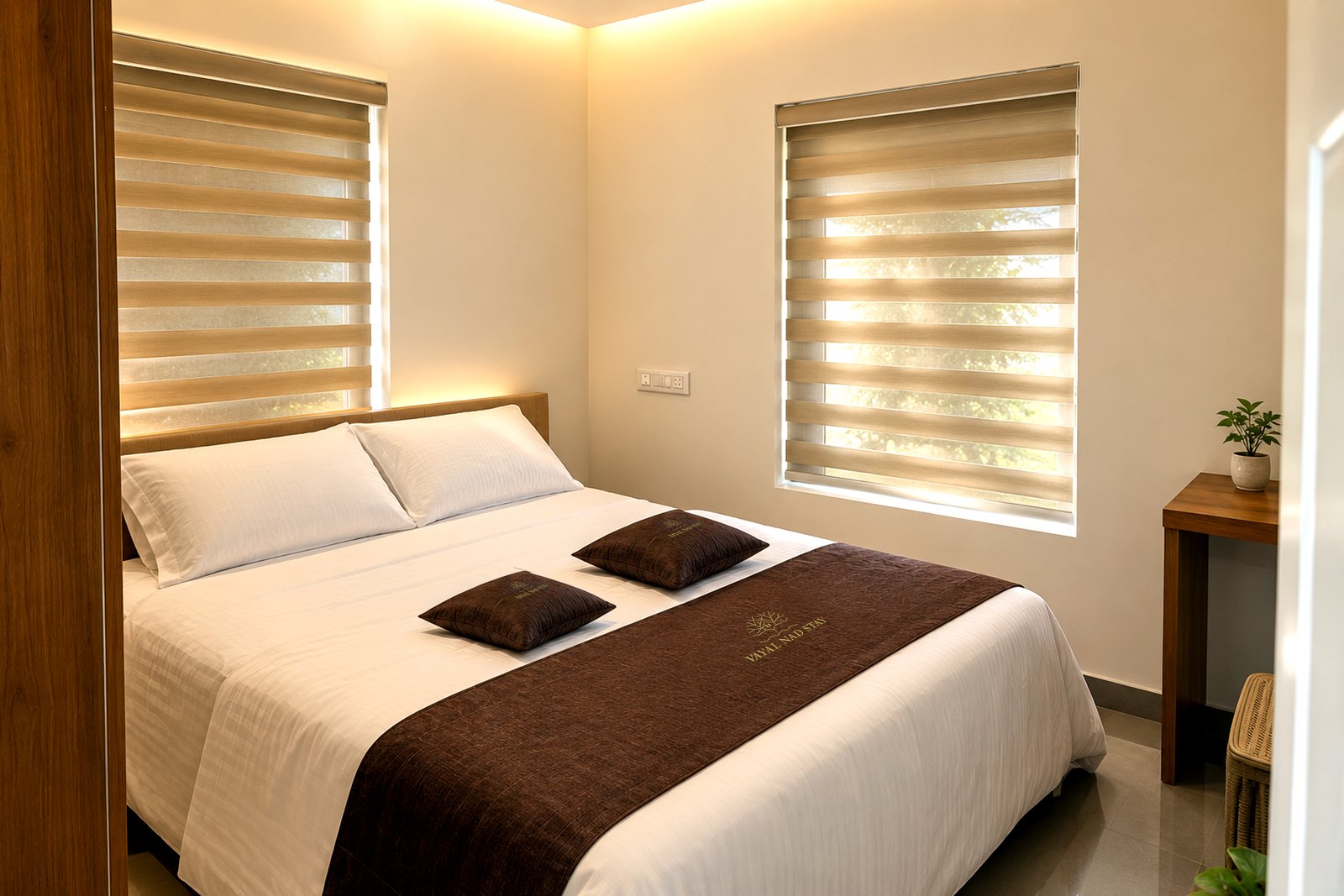Bedroom with a large bed, brown runner and branded cushions between two windows with layered blinds in the Paddy Field Balcony Room